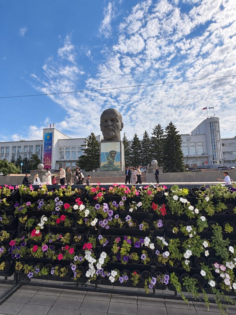 Ulan-Ude Sovyet Meydanı’ndaki dev Lenin başı anıtı, çevredeki insanlar ve ön plandaki çiçekler